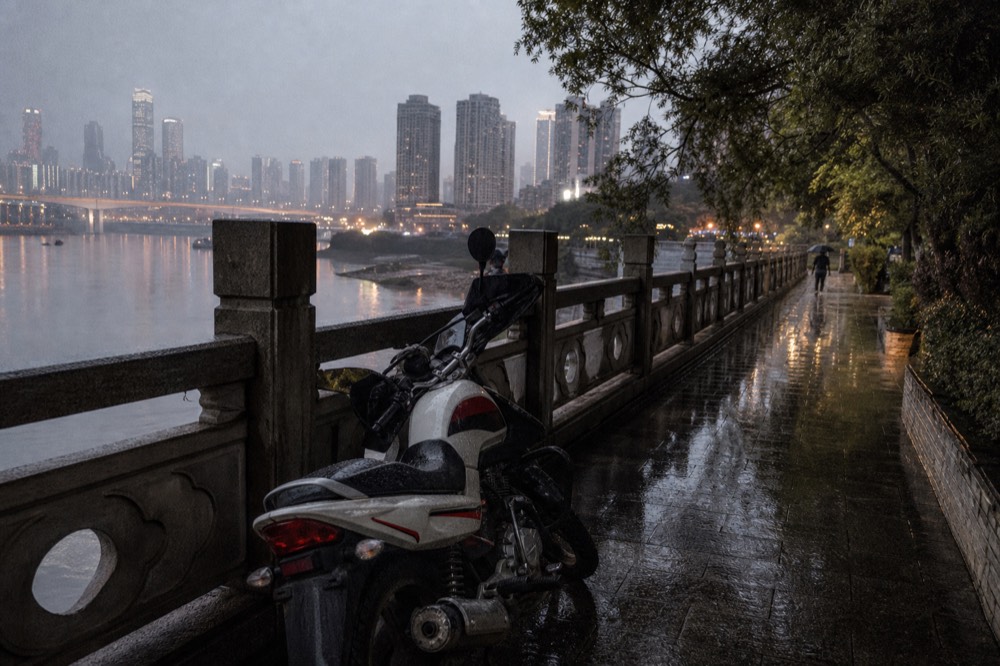 A motorcycle parked beside a lit riverside promenade at night with tower blocks glowing across the river