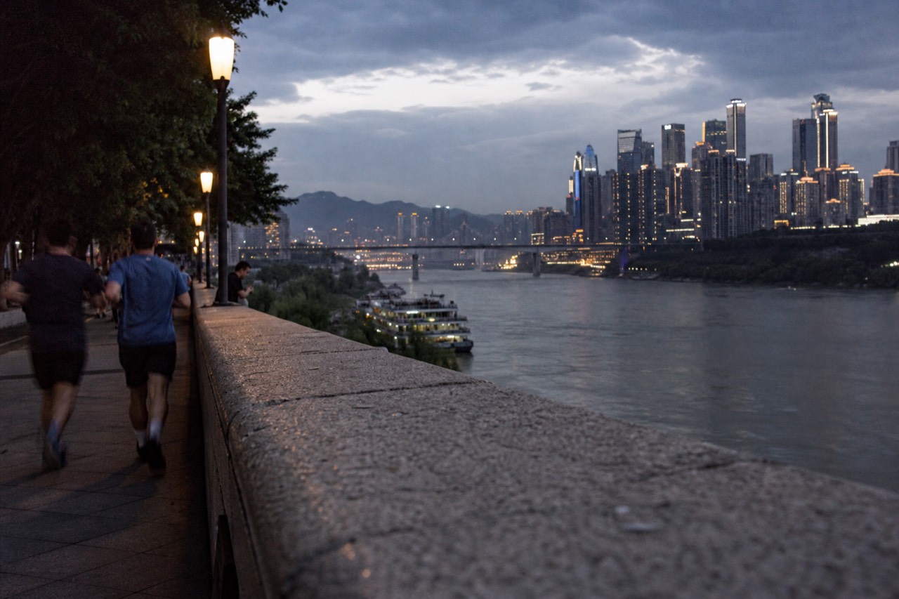 Illuminated skyline towers and a bridge over a wide river seen from a dark riverside promenade