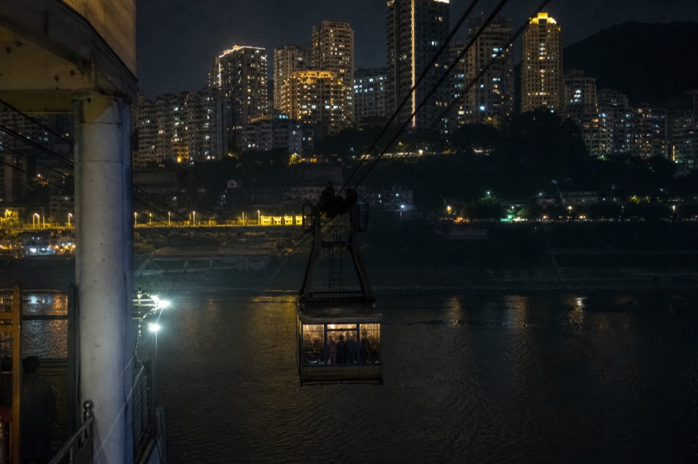 A cable car cabin crossing a dark river toward a hillside of illuminated apartment towers
