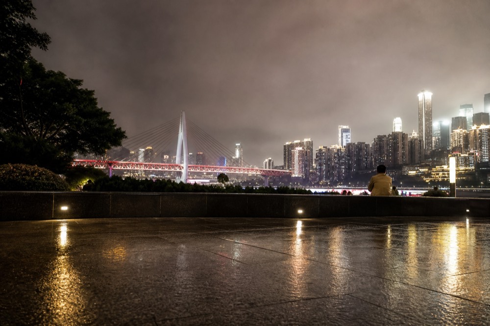 A riverside plaza at night with illuminated towers and a cable-stayed bridge across the river