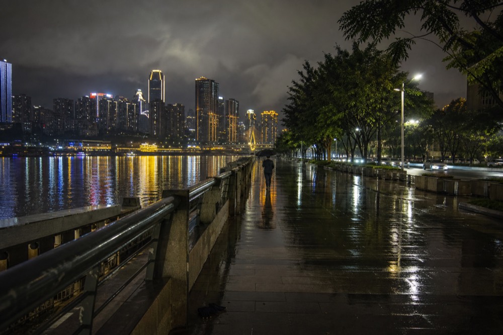 A riverside promenade at night with lit towers on the far shore and wet pavement reflecting the lights