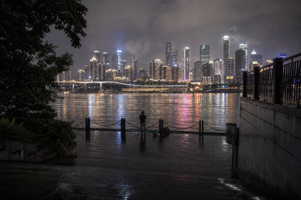 Neon-lit high-rise buildings reflected in a wide river at night, seen from a dark riverside promenade