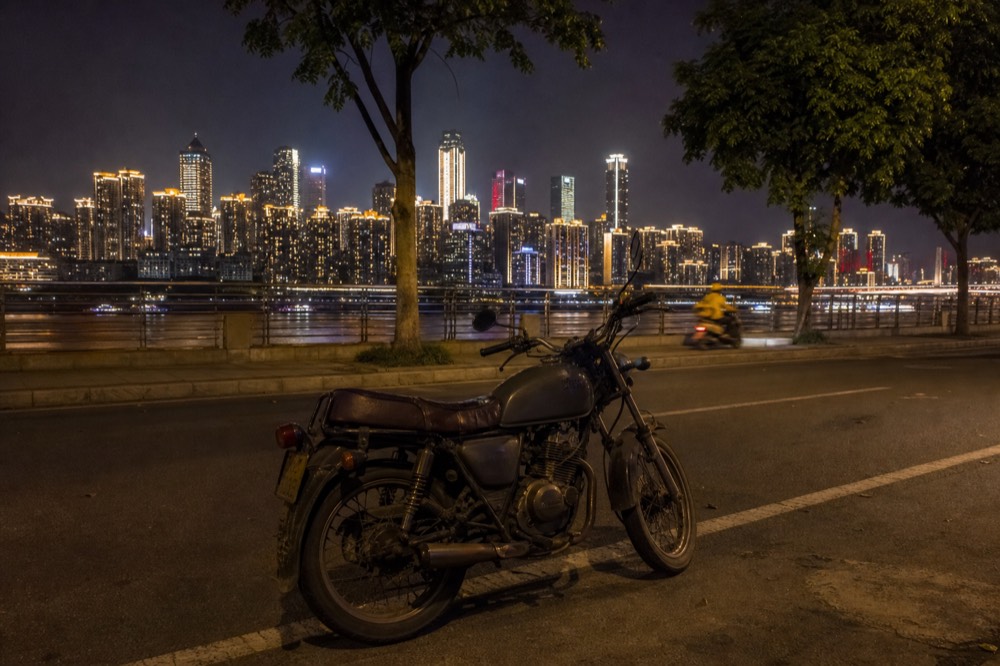 A parked motorcycle on a riverside road at night with a brightly lit city skyline in the background