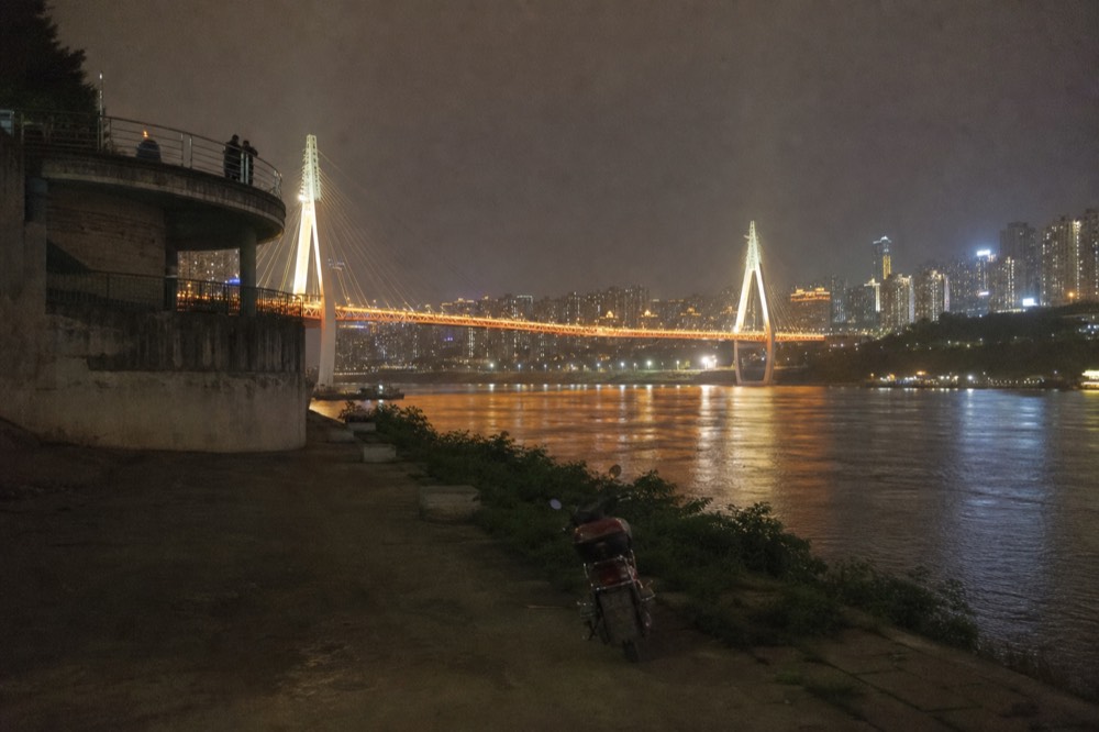 Illuminated riverside promenade with lit bridge towers reflected in the Yangtze at night