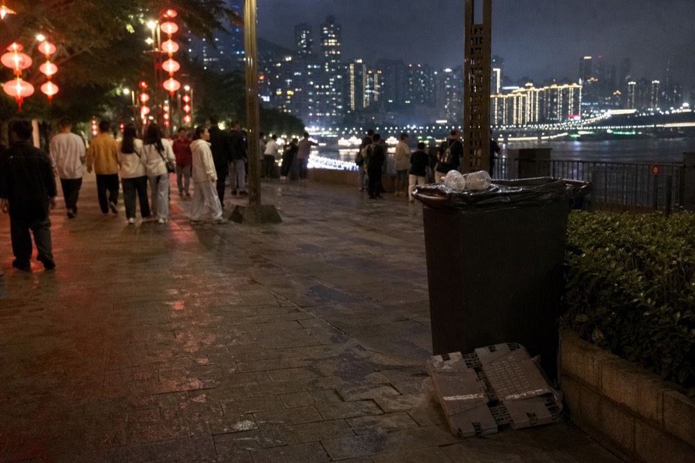 Crowded riverside walkway at night with city towers glowing across the water