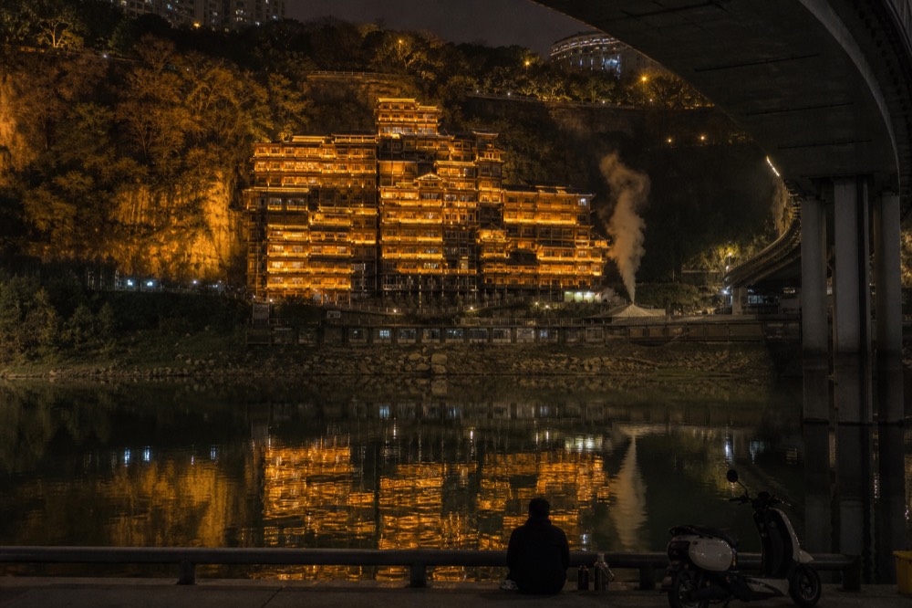 Illuminated stilt-house complex built into a cliff above a river, reflecting in the dark water