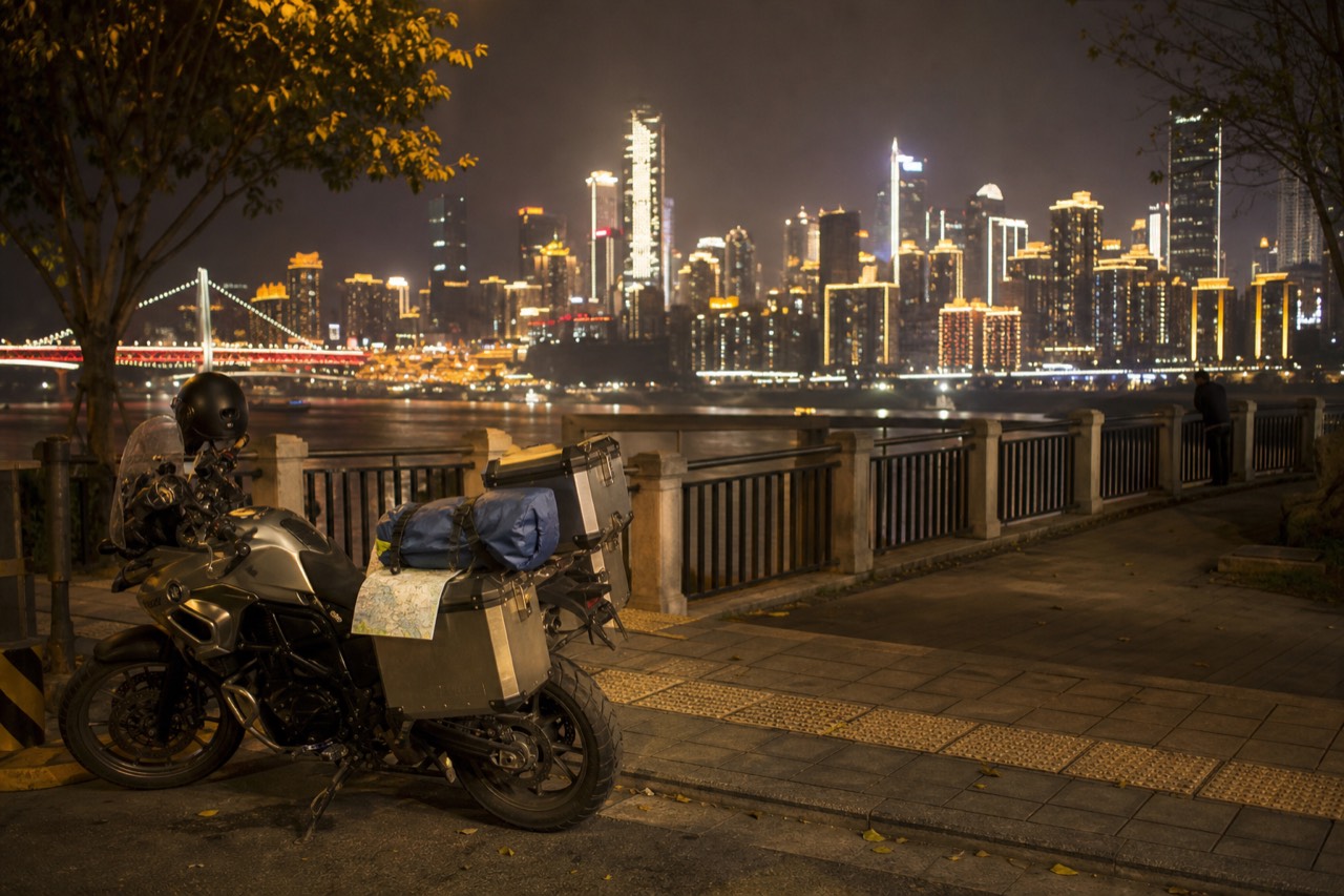 A parked motorcycle beside a lit riverside promenade with a city skyline of towers and bridges across the water at night