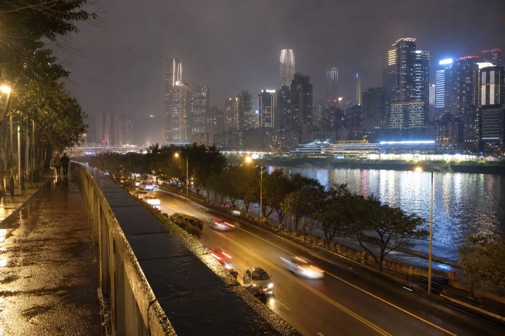 A lit urban riverfront road at night with illuminated towers reflected across the water