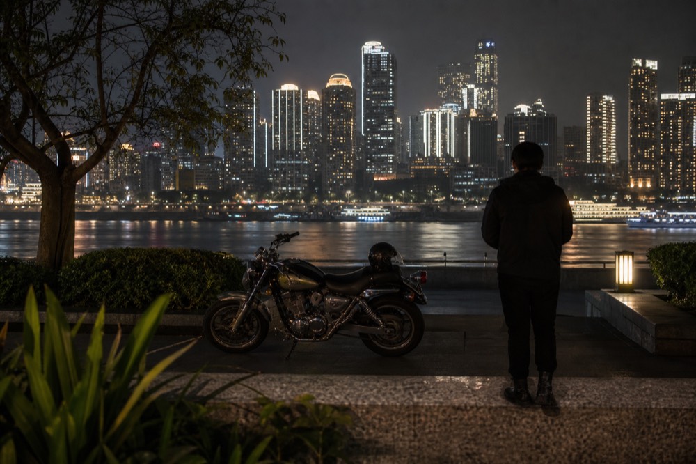 Motorcycle and rider waiting on a riverside road at night with skyline lights