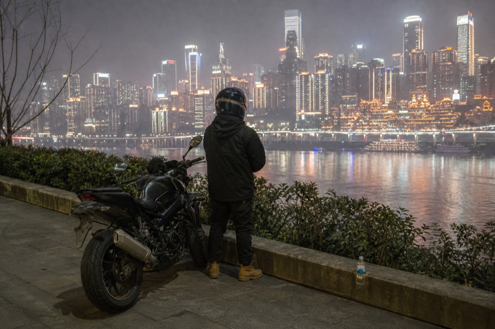 Person in a helmet posing beside a parked motorcycle with city lights in the background