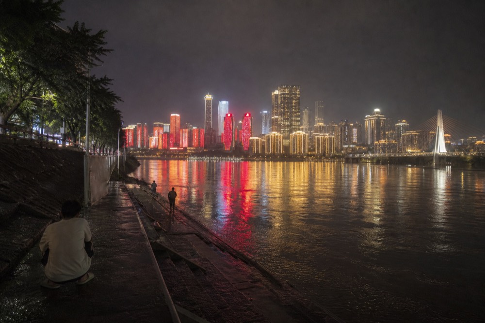 Nighttime riverside road with illuminated towers reflected in the river and a bridge in the distance