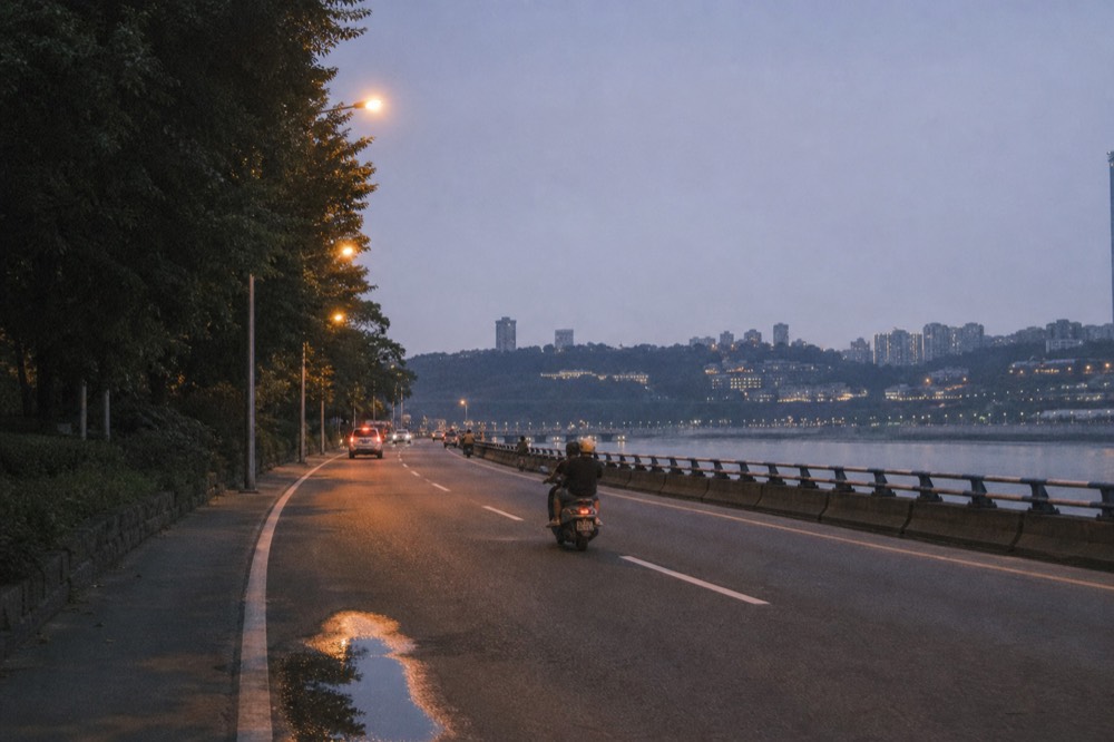 Rider and passenger on a motorcycle moving along a riverside boulevard at dusk