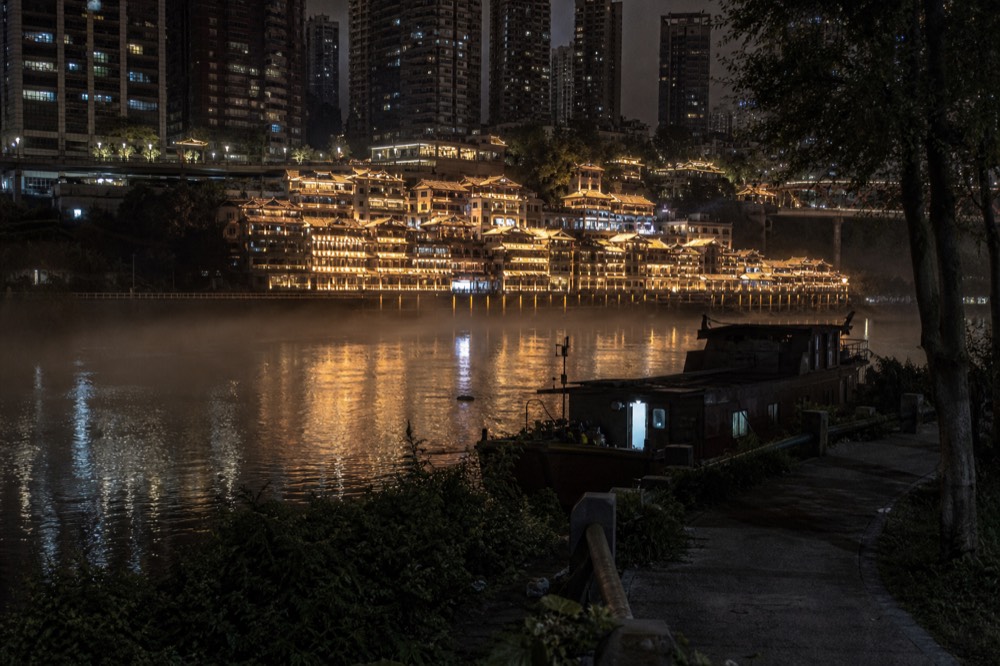 Night view across a wide river toward a dense illuminated city skyline with a brightly lit stilt-house complex at the waterline.