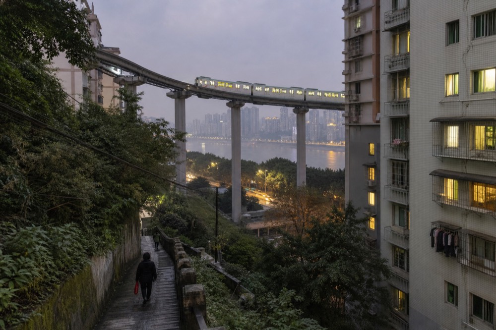 Elevated metro viaduct curving between high-rise apartment towers in a dense hillside city at dusk.