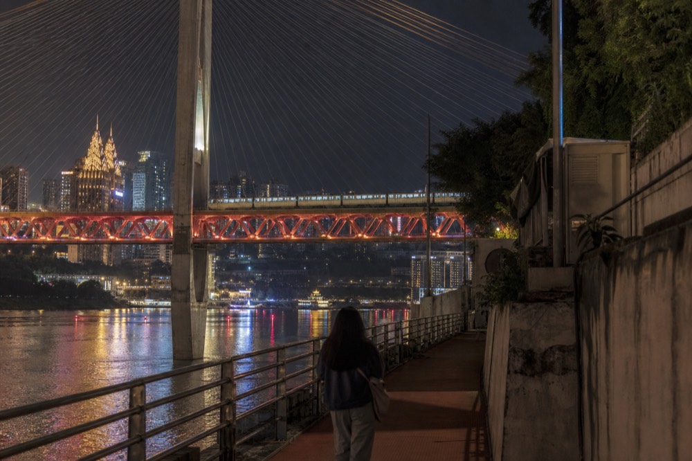 Night view of a monorail train crossing an elevated bridge between brightly lit towers above a river