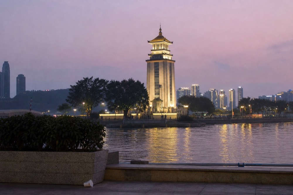 Plaza by a river at early evening with a bell tower structure and city lights reflecting on the water