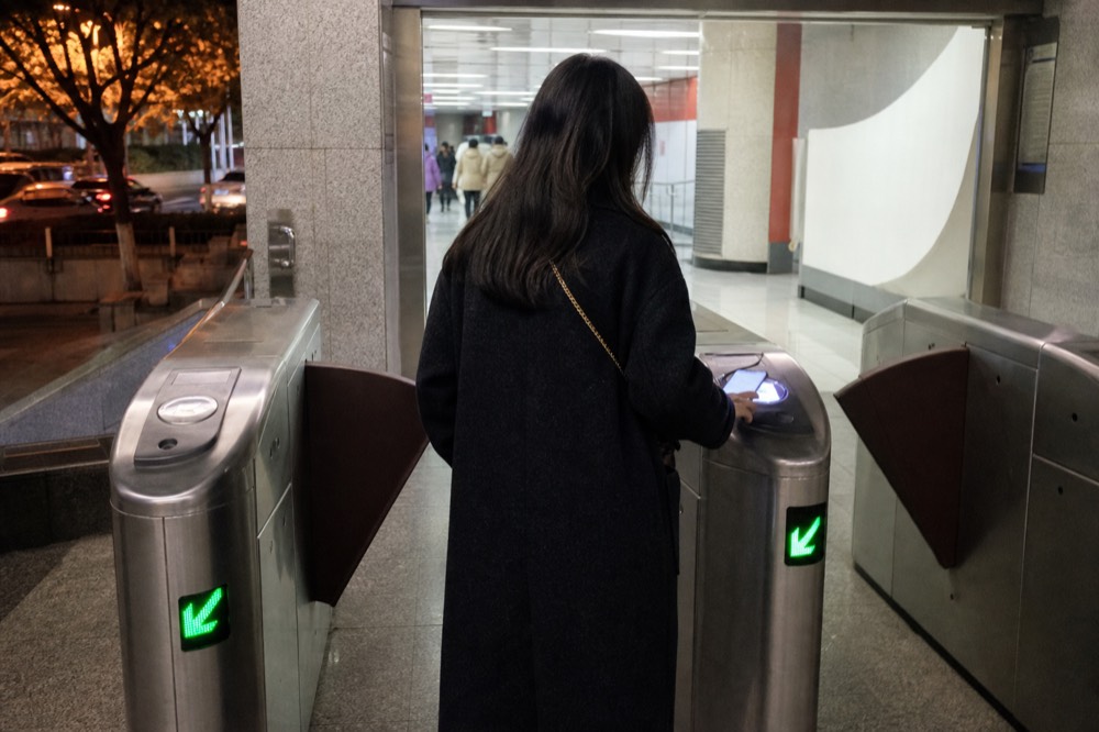 Rider seen from behind holding a glowing phone at a metro station fare gate at night