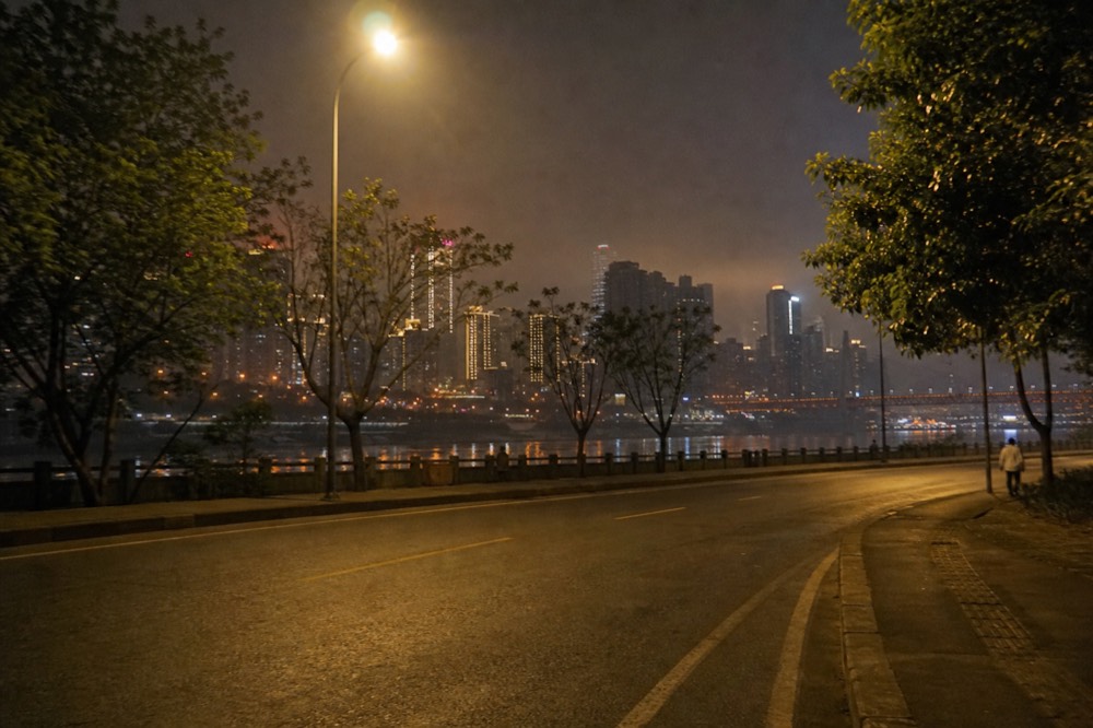 Two-lane riverside road at night with the illuminated towers of the opposite bank rising above the water
