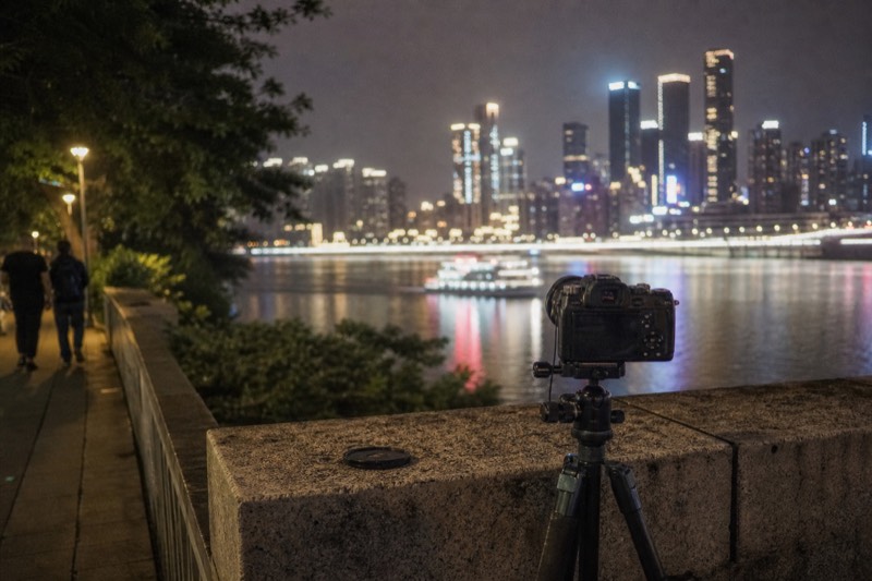Camera on a tripod at the Chongqing waterfront after dark, framed toward the far bank, where the lit skyline sits soft and out of focus behind the lens.